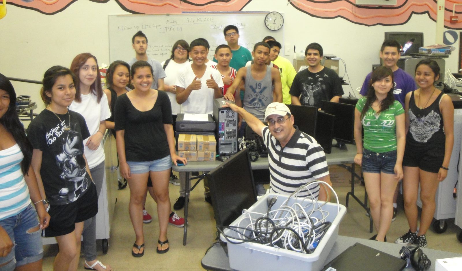 Students crowded around a table stacked with donated computers, monitors and cables, smiling and giving thumbs up